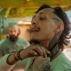 Ein Mann mit Schmuck und Tattoos hält eine Klinge an seinen Hals; ein anderer Mann ist unscharf im Hintergrund.