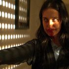 Keine sichtbaren Texte. Eine Person mit langem Haar, blutverschmiertem Mund, steht im Licht, das durch ein Fenster fällt.