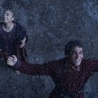 Zwei Menschen schauen lächelnd nach oben im Schneefall, Arme ausgebreitet. Eine freudige Szene.