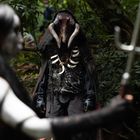 Eine maskierte Gestalt in einem düsteren Wald, mit knochigen Rüstungselementen. Im Vordergrund eine Figur mit Speer.