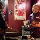 "Open"-Leuchtschild. Zwei Personen, eine am Verkaufstresen, die andere hält VHS. Wandposter mit "DAY". Gemütliches Ambiente.