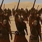 Eine Gruppe vermummter Gestalten steht in einer Wüste, hebt Waffen gen Himmel.