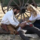 Zwei Personen kümmern sich um einen verletzten Mann auf dem Boden vor einem Wagenrad. Ihre Hände sind blutverschmiert.