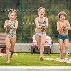 Drei lachende Kinder im Badeanzug laufen auf Gras, jedes hält ein kleines Entenküken. Im Hintergrund Liegestühle und Handtücher.