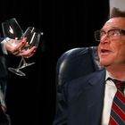 Eine Frau in Uniform hält Wein und Gläser bereit. Gegenüber sitzt ein lächelnder Mann im Anzug.