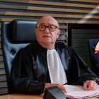 Ein älterer Mann in einer schwarzen Robe sitzt an einem Tisch, darauf ein Mikrofon und ein Stapel Papiere.