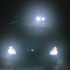 Zwei Fahrzeuge mit eingeschalteten Scheinwerfern auf dunkler Straße in der Nacht, das vordere im Vordergrund.