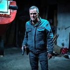 Text: "DISCO M". Ein älterer Mann in blauer Jacke steht in einer dunklen Umgebung, hält eine Pistole und wirkt angespannt.