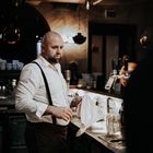 Ein Barkeeper mit Glatze und Bart poliert Gläser an einer Theke; er trägt ein weißes Hemd mit Hosenträgern.