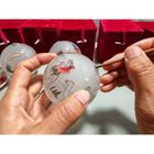 Eine Hand bemalt eine Weihnachtskugel mit Szenen, darunter Reiter auf Kamelen. Im Hintergrund sind rote Samtboxen.