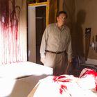 Ein Mann steht in einem Raum mit blutbespritzten Wänden und Messern an der Wand. Ein Rollstuhl ist im Hintergrund.