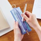 Eine Hand hält einen weißen Stift über ein lilanes Federetui mit Reißverschluss auf einem Holztisch.