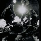 Drei Männer beugen sich über eine am Boden liegende Person, beleuchtet von starkem Licht.