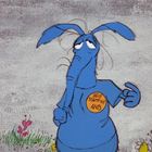 Auf einem Button steht "HELP STAMP OUT ANTS". Ein blaue, cartoonartige Ameisenbärfigur trägt ihn. Gras und Blumen sind zu sehen.