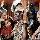 Drei Personen in traditioneller Kleidung mit Federschmuck, ernster Ausdruck, sitzen oder stehen nebeneinander.