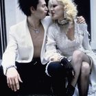 Ich kann keine Texte oder Namen erkennen. 

Ein Paar in kontrastreicher Kleidung sitzt, er in einem weißen Blazer, sie in Spitzenkleid und mit extravaganten Accessoires.