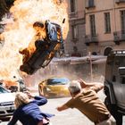 Ein brennendes Auto fliegt durch die Luft. Menschen laufen weg, ein goldenes Auto mit "CAR SICK" Kennzeichen. Gebäude im Hintergrund.