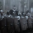 Römische Soldaten in Rüstung mit Schilden und Speeren stehen im Wald, begleitet von zwei Reitern.