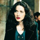 Eine Frau mit dunklem Haar und rotem Lippenstift trägt eine blaue Bluse und schwarzen Blazer.