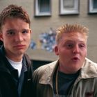 Zwei junge Männer stehen draußen; der Hintergrund zeigt ein Gebäude mit Fenstern.