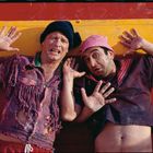 Zwei Männer in zerrissener Kleidung, mit erstauntem Gesichtsausdruck, lehnen an einer rot-gelben Wand.