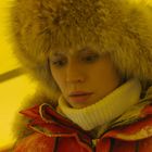 Kein Text. Eine Person mit Pelzhut und rotem Mantel in einem gelben Zelt, fokussiert und nachdenklich schauend. Schnee sichtbar.