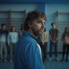 Ein Mann in blauer Jacke im Vordergrund, schaut zur Seite. Im Hintergrund stehen unscharf mehrere Jugendliche.