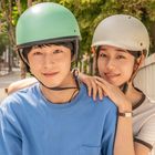 Zwei Personen auf einem Roller, beide tragen Helme. Eine Person im blauen Shirt, die andere umarmt sie von hinten. Straße im Hintergrund.