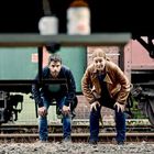 Zwei Personen in gebückter Haltung schauen unter einem Objekt hindurch, umgeben von Eisenbahngleisen und Waggons.