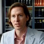 Ein Mann mit braunen Haaren trägt ein gestreiftes Jackett vor Regalen mit vielen Büchern.