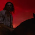 Ein Mann mit langen Haaren reitet auf einem Pferd vor einem dramatisch roten Himmel.