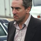 Ein Mann mit ernstem Gesichtsausdruck und zurückgekämmtem Haar trägt ein Hemd und ein Jackett im Freien.