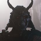 Ein dunkler Krieger mit markanter Rüstung, leuchtend roten Augen und großen, gebogenen Hörnern auf dem Helm.