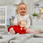 Ein lächelndes Baby sitzt auf einer Decke, hält ein Kuscheltier und trägt rosa Kleidung. Im Hintergrund ist ein Kinderzimmer.