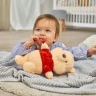 Ein Baby liegt auf einer Decke und knabbert an einem roten Spielzeug, während es einen Plüschbären hält. Im Hintergrund steht ein Kinderbett.