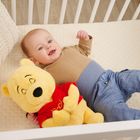 Ein Baby liegt in einem Bett und hält ein gelbes Kuscheltier mit rotem Shirt in den Armen.