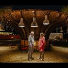 Ein Mann und eine Frau stehen unter einer beleuchteten, pilzförmigen Struktur bei Nacht, umgeben von Lichtern.