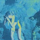 Keine Texte zu sehen. Eine Person mit Kamera am Strand von Kreatur in Schuppenanzug verfolgt. Szene wirkt inszeniert.