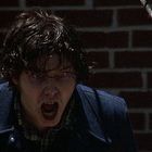 Kein Text erkennbar. Eine Person mit weit geöffnetem Mund und nassen Haaren steht vor einer roten Ziegelwand.