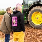 Ein Mann mit pinkem Irokesenschnitt trägt einen schwarzen Hoodie mit der Aufschrift "SPUD MAN". Daneben ein Mann. Im Hintergrund steht ein Traktor.