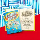 Text: "SCUBA CATS", "BOOK 1". Illustration: Zwei fröhliche Katzen in Taucheranzügen mit einem Fisch auf einer blauen und roten Fläche.