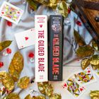 Zwei Bücher mit "The Gilded Blade" an den Buchrücken, umgeben von Spielkarten und goldenen Blättern auf Stoff.