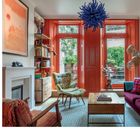 Text über Inneneinrichtung. Raum mit orangen Türen, bunter Sessel, lila Sofa, Pflanzen, und auffälliger blauer Deckenlampe.