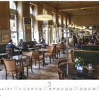 "Café Westend", darunter Kalenderzahlen. Eleganter Kaffeehausraum mit Holzstühlen, runden Tischen und Kronleuchtern.
