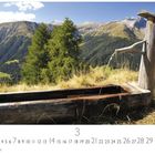 Alpenbrunnen, Märzkalender, Berglandschaft mit einem alten Holzbrunnen und fließendem Wasser, umgeben von Wiesen und Tannen.