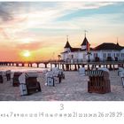 "Seebrücke Ahlbeck" und "CASARES EDITION". Eine Seebrücke mit Gebäude bei Sonnenuntergang, vorne Strandkörbe am Sandstrand.