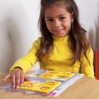Ein Mädchen in gelbem Pullover spielt mit gelben Karten mit roten Buchstaben darauf an einem Holztisch.