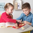 Zwei Kinder spielen und lernen an einem Tisch mit einem Buch und Zahlenplättchen. Ein Regal mit Deko ist im Hintergrund.