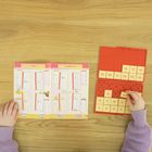 Ein Kinder-Rechenbuch mit Zahlen und Aufgaben. Rechts ein rotes Zahlenfeld mit beige Zahlenplättchen. Hände sind sichtbar.