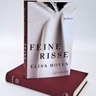 Ein Buch mit dem Titel "Feine Risse" von Elisa Hoven, bezeichnet als "Roman", steht auf einem braunen Buch.
